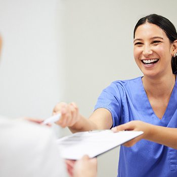 a smiling woman in scrubs handing a patient forms on a clipboard